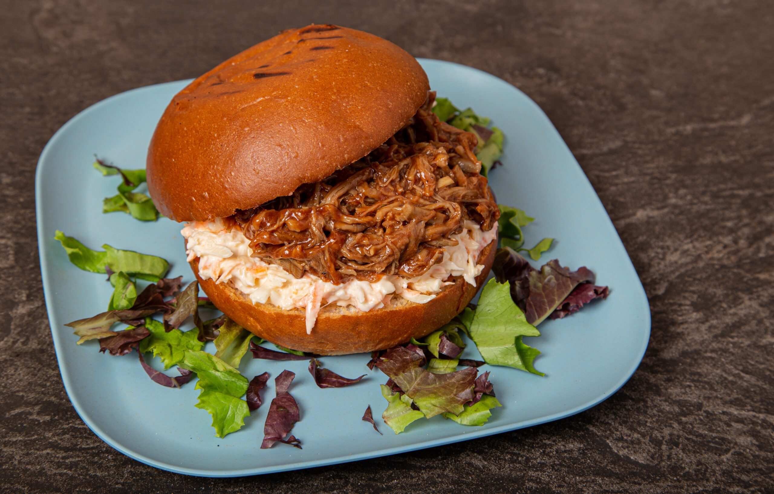 A closeup shot of an open juicy pulled meat burger surrounded by greens on a blue plate